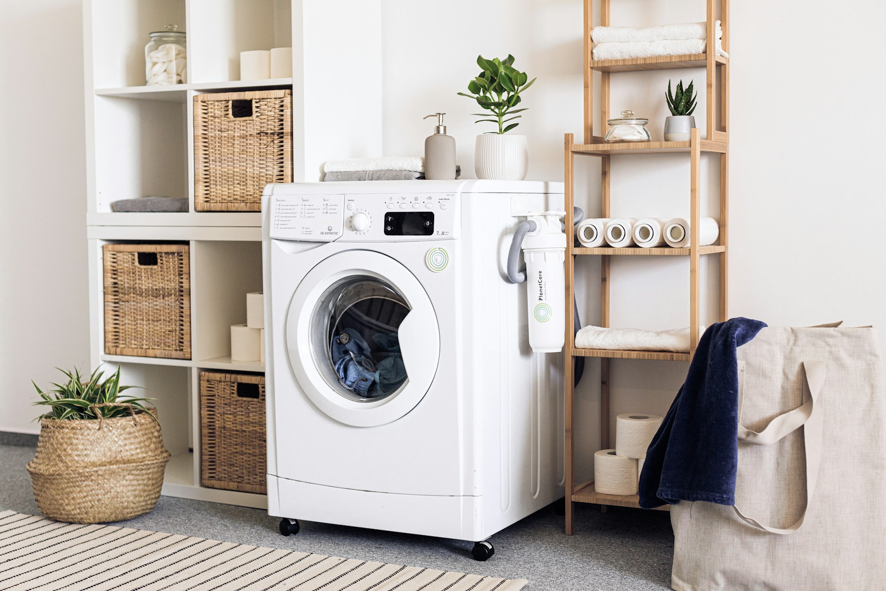 A modern white front-loading washing machine in a bright laundry room with white shelving, baskets, towels and plants