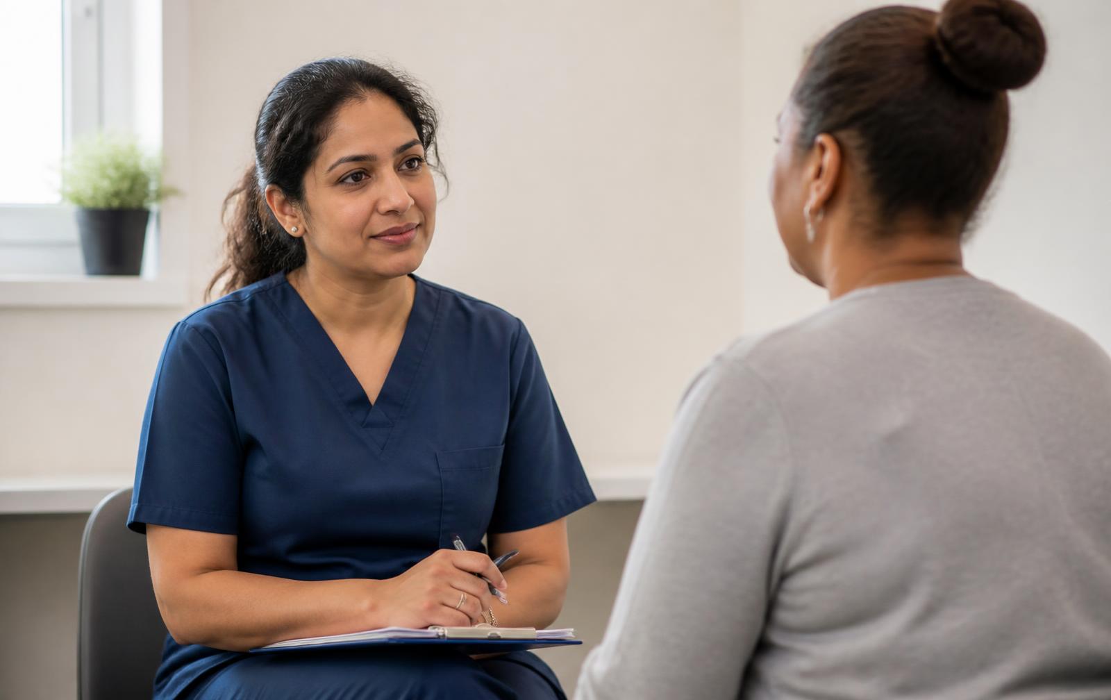 A healthcare professional in navy scrubs listens attentively to an adult patient in a bright clinic room.