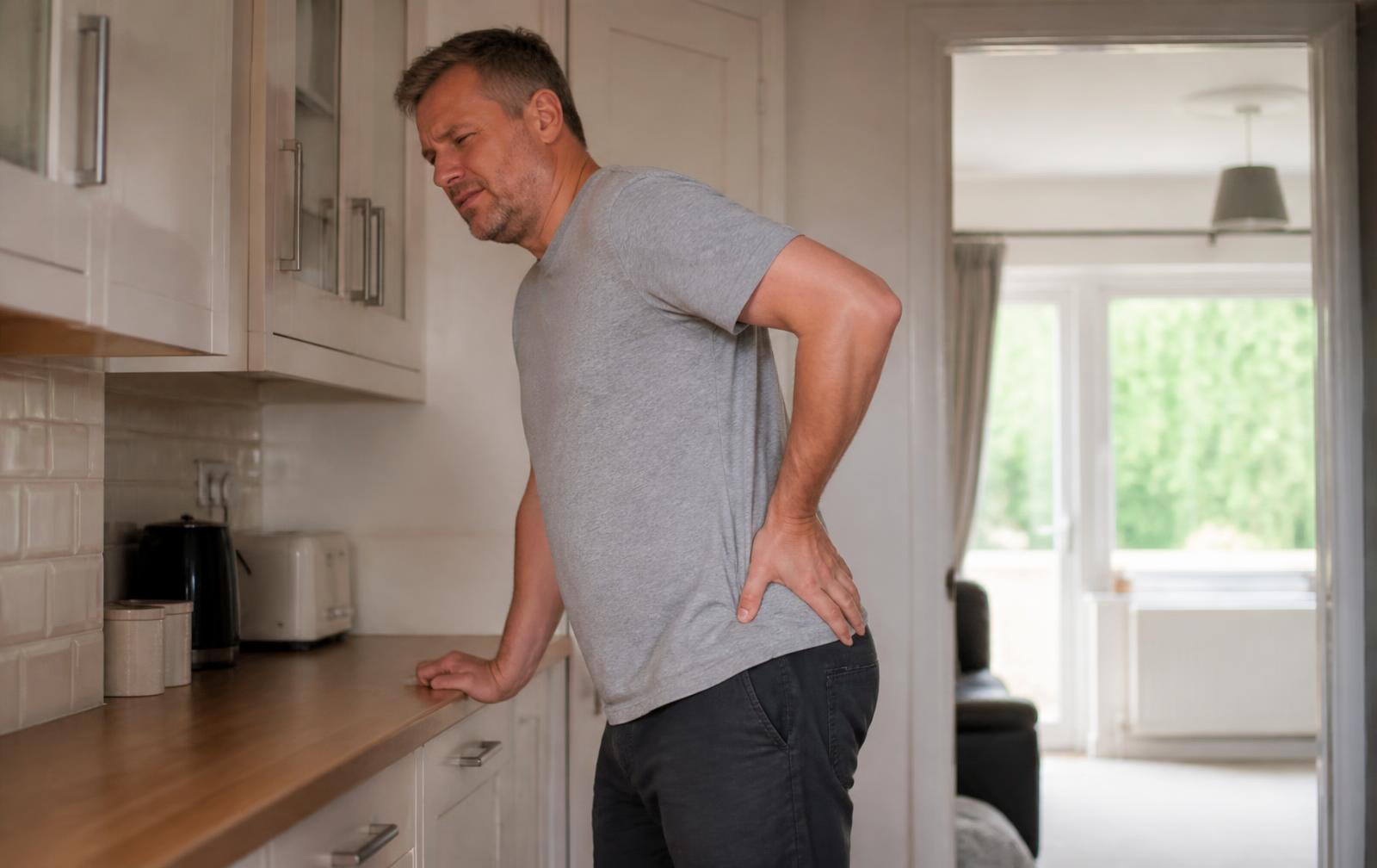 A fully clothed man in his forties standing in a kitchen, resting one hand on his lower back and buttock.